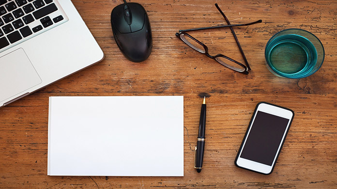 A laptop, mouse, glasses, paper, pen, phone, and cup on a table